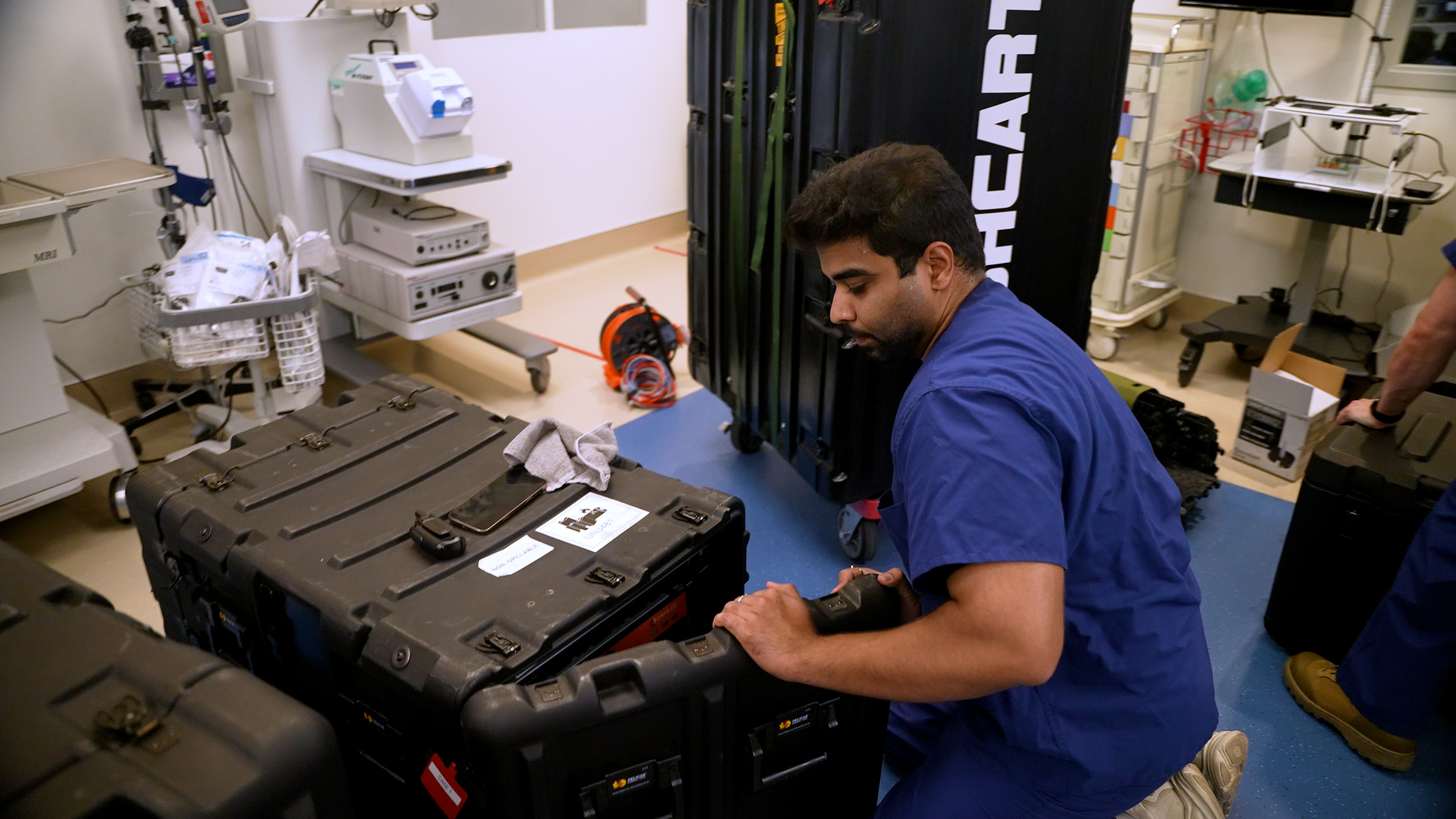 A man wearing blue scrubs kneels on the ground in a hospital room to open a large black container.